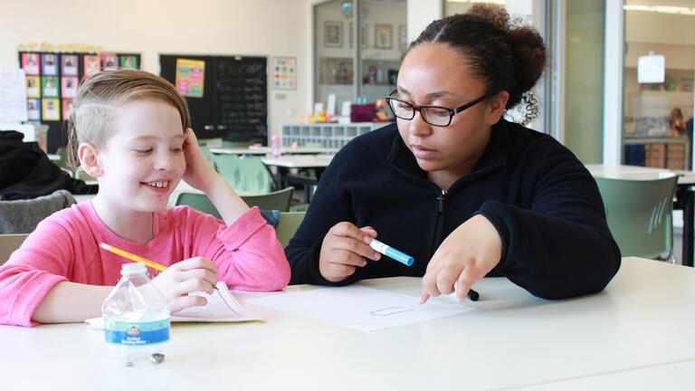 Teacher sits with child at desk writing on paper with pencils in hand