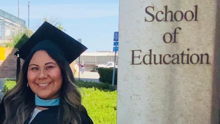 Carmen Candelaria MA '20 stands by a sign reading "School of Education"