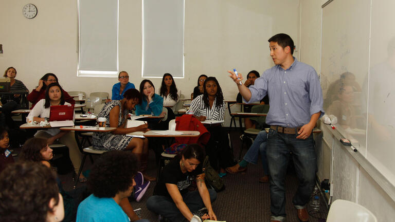 Students sit at desk and watch teacher at white board