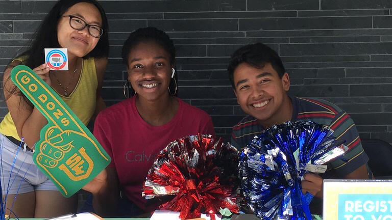 Three students run a voter registration table hold up pom-poms, a foam finger, and an I Voted sticker.