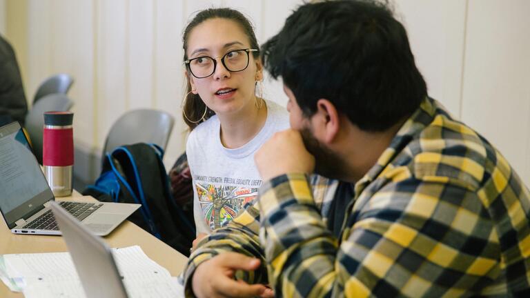 Two students sit as desk and discuss