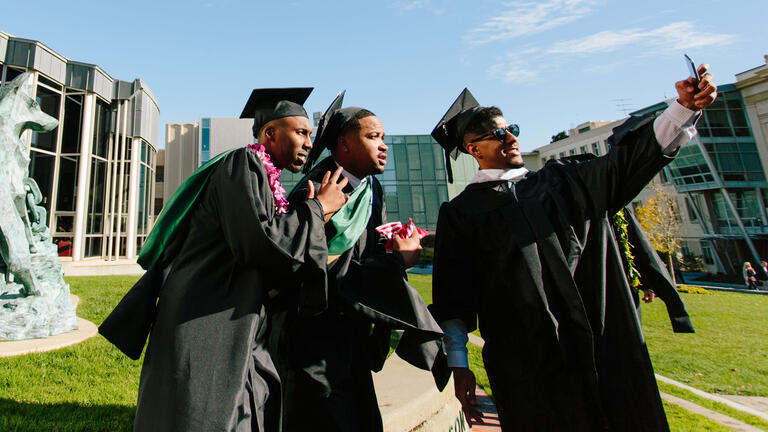 Three graduates in regalia take a selfie in front of the library.