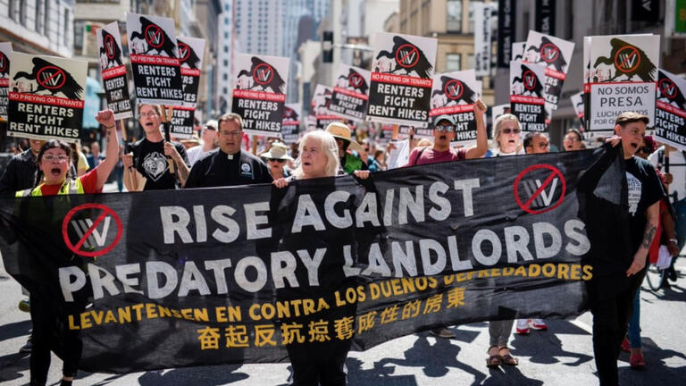 Protestors march carrying a banner that says "Rise Against Predatory Landlords"