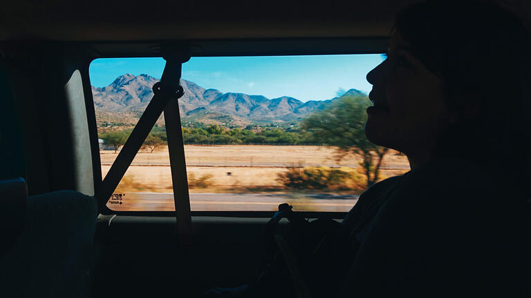 Car passenger silhouetted in shadow with passing mountains and desert landscape framed by window