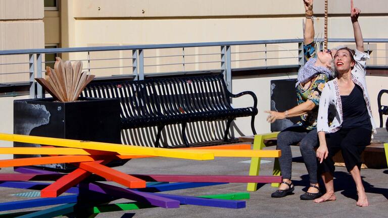 Two people pointing at the sky while sitting on a bench on a rooftop.