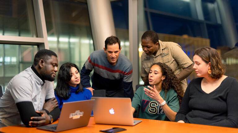 students stand over laptops in class