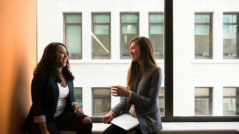 Two women talking in a room in front of a window