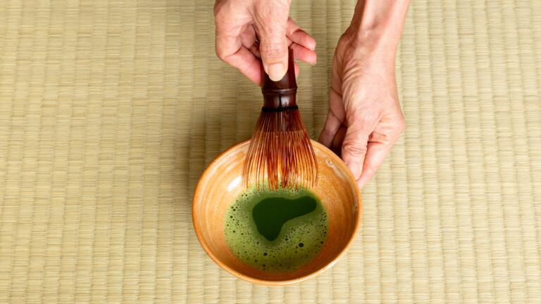 Hands holding a cup of tea with a whisk over a tatami mat