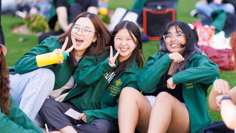 3 students smiling, sitting on the grass wearing green USF jackets
