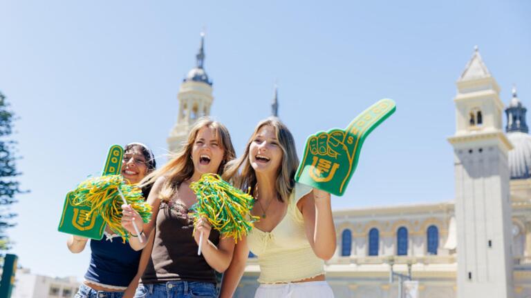 3 students cheering while holding USF foam fingers and green and gold pom-poms