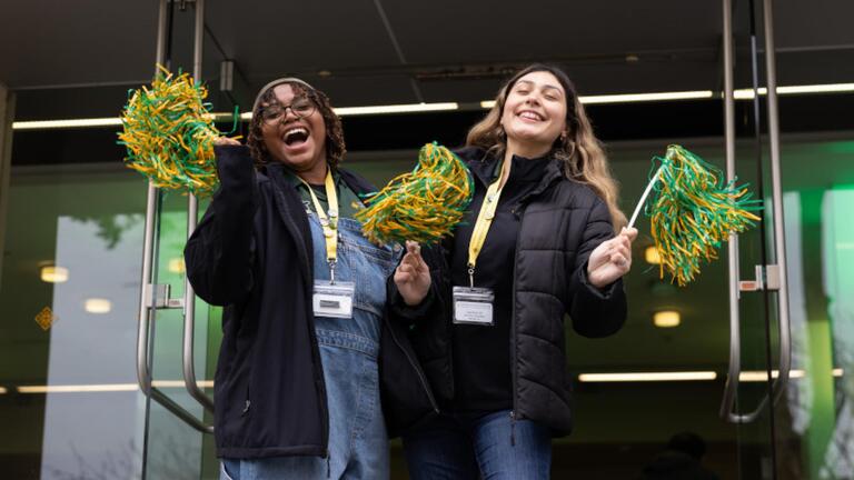 Two students cheering and waiving green and gold pom-poms.