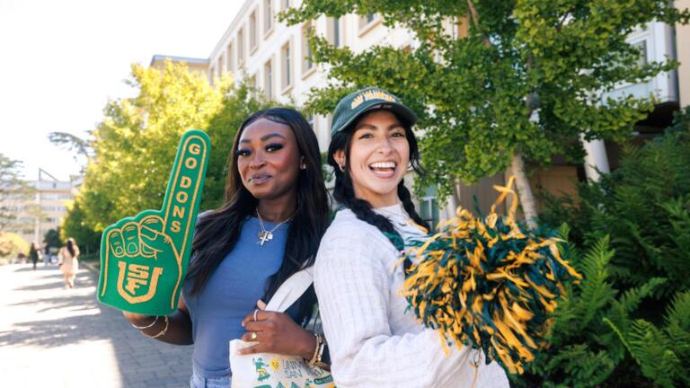 Two students standing back to back holding a USF foam finger and green and gold pom-poms.