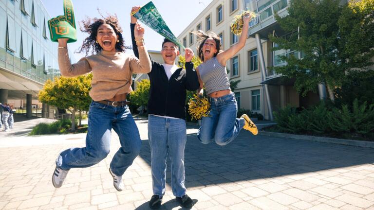 3 Students Jumping in the air holding a USF foam finger, a pendant and green and gold pom-poms