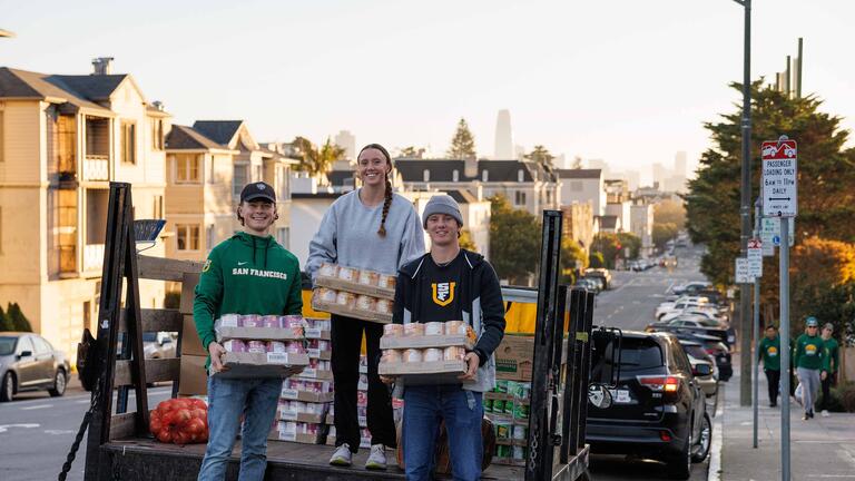 Three USF students on a truck bed holding cases of canned food items to be delivered