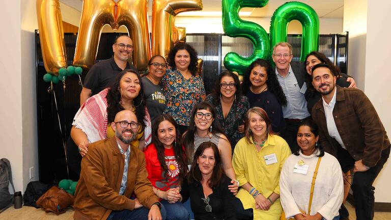 Two rows of people huddled for a photo with "IME 50" balloons behind them