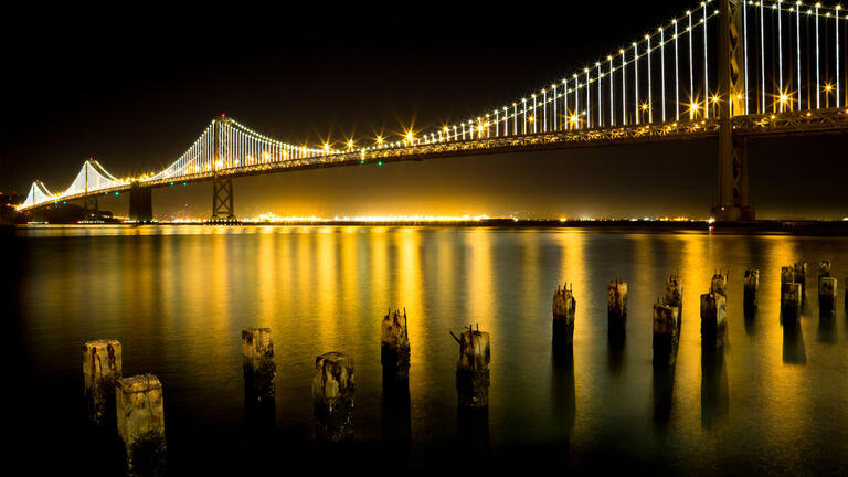 Bay Bridge lit up at night, golden light reflecting on the bay