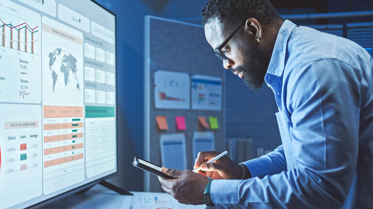 Man in business attire looking at phone and monitor screen, which displays data
