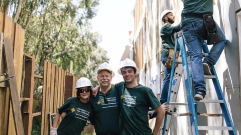 three people standing with hard hats on smiling and two people working on a ladder