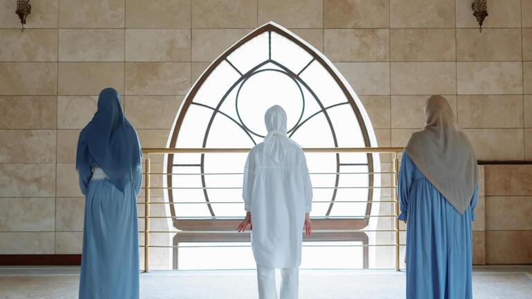 Three women wearing headscarves/hijabs facing the wall with a window