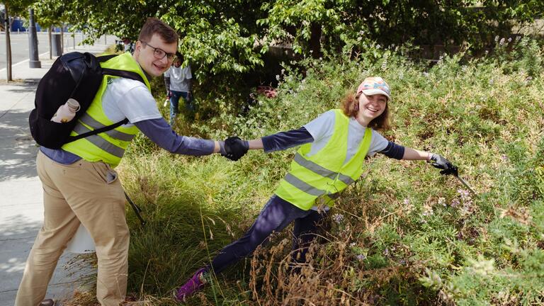 Two people in safety vests hold hands while picking up litter near bushes, with one leaning forward and smiling. Another person works in the background.