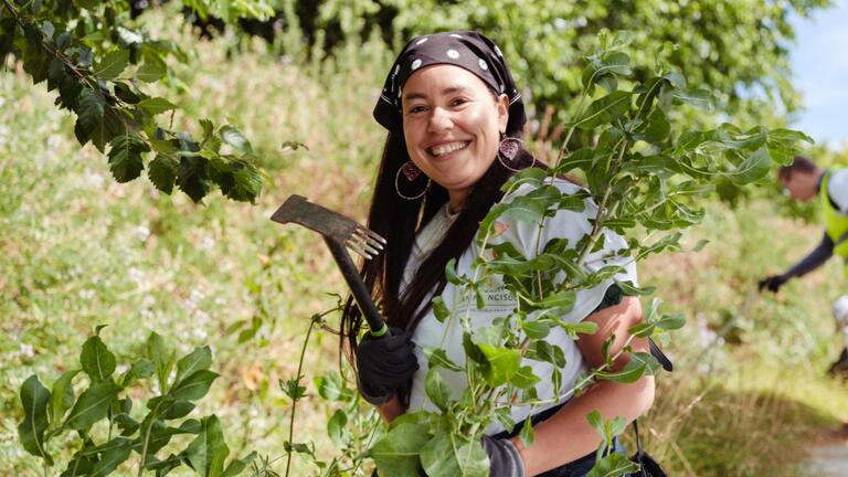 Woman wearing gloves and a bandana smiles while holding a gardening tool and standing among green plants outdoors.