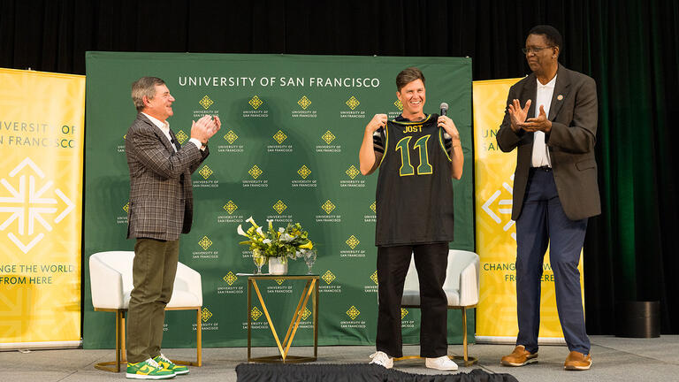 Colin Jost on stage holding up a USF basketball jersey with Jeff Silk ’80 and Bill Cartwright holding