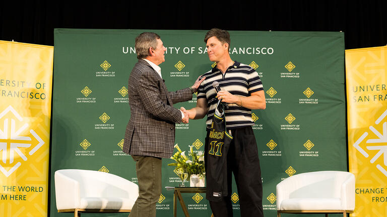 Colin Jost shaking hands with Jeff Silk ’80 and holding a USF basketball jersey on stage at the Silk Speaker Series