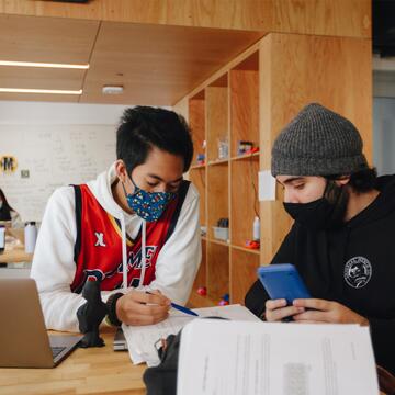 Student holding pen discusses project with another student holding phone while sitting at a desk