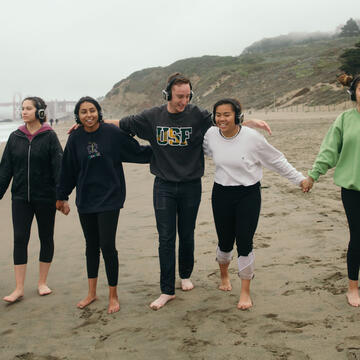 A group of students holding hands walk down Baker Beach in SF.