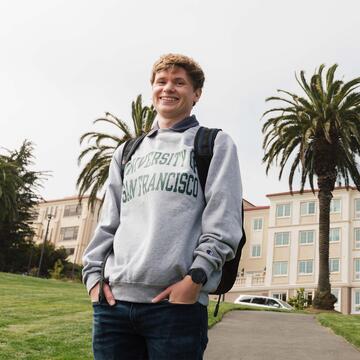 Student in USF sweatshirt stands in front of the Lone Mountain dorms.