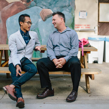Two people sit on a bench in front of a mural and talk