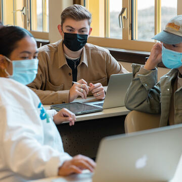 Three students talk in a group while sitting in front of open laptop computers