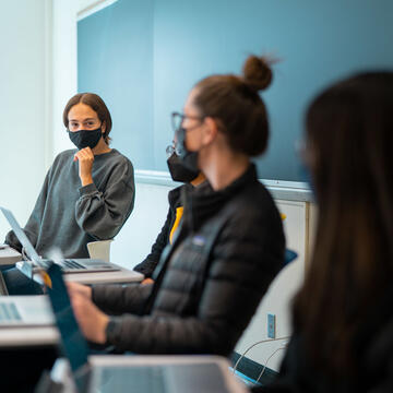 Four student with laptops sit at desks and talk in front of blackboard
