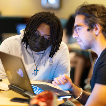 Two students sit at a table and collaborate in front of an open laptop compute