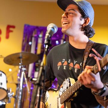 Student playing a guitar sings into a microphone in front of a hand made sign that reads KUSF