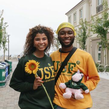 Two students holding flowers and a stuffed animal pose outside the dorms.