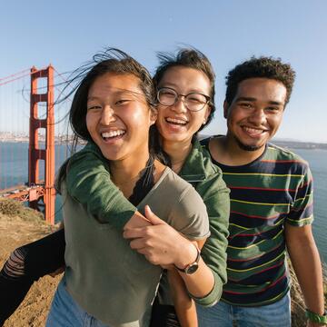 Three students embrace and smile on a hilltop overlooking the Golden Gate Bridge.