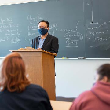 Literature instructor at the podium stands in front of black board that has explanations of a book's main characters on it