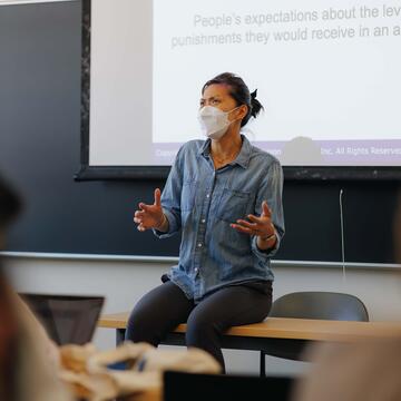 Professor lectures in front of a slideshow while sitting on a table