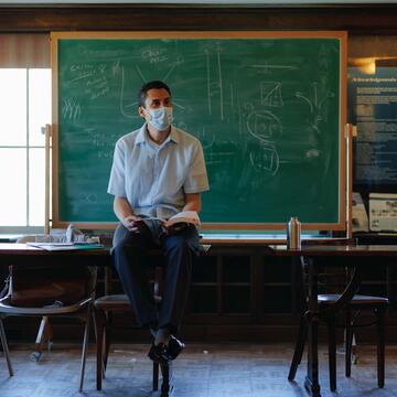 Instructor sits on a desk lecturing in front of a chalkboard