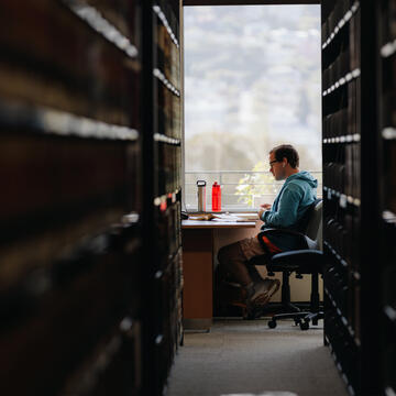 Student seen through a row of bookshelves studies at a table.