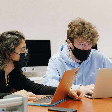 Two students look at something on a laptop while in class.