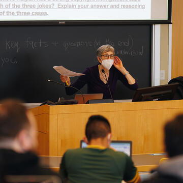 Professor who is in front of a black board and a screen stands at the podium with left hand to their ear and the other holding papers