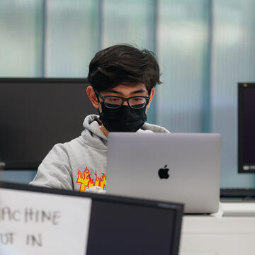 Student sits in a computer lab working on a laptop while surrounded by monitors