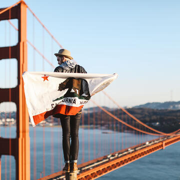 Student wrapped in a California flag stands on a high point with the Golden Gate Bridge behind.