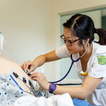 nursing student in scrubs holds stethoscope to manikin's chest