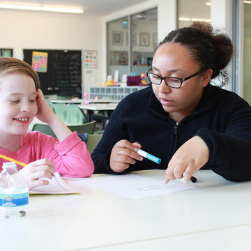 Teacher sits with child at desk writing on paper with pencils in hand