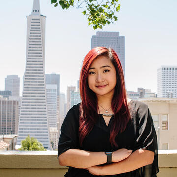 Student stands cross armed on a balcony with SF downtown skyline behind.