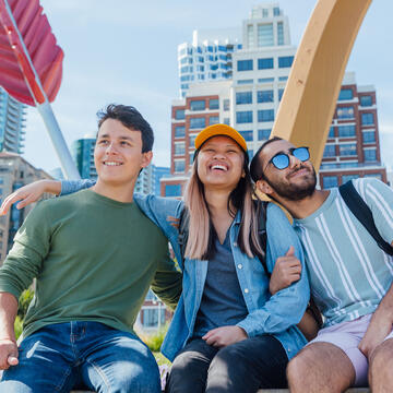 Three students sit with arms around each other in front of bow and arrow sculpture downtown.