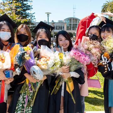 Six international students pose holding flower bouquets and wearing graduation regalia